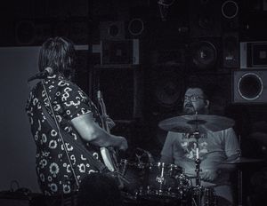 Musicians perform on stage in a dimly lit venue with a guitarist in a patterned shirt and drummer behind a kit.