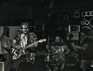 Black and white photo of a band performing on stage with a guitarist in a patterned shirt and drummer in background.