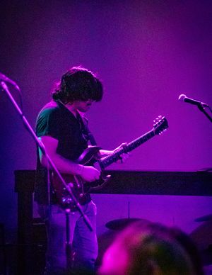 Guitar player performing on stage bathed in vibrant purple lighting during a live concert performance.