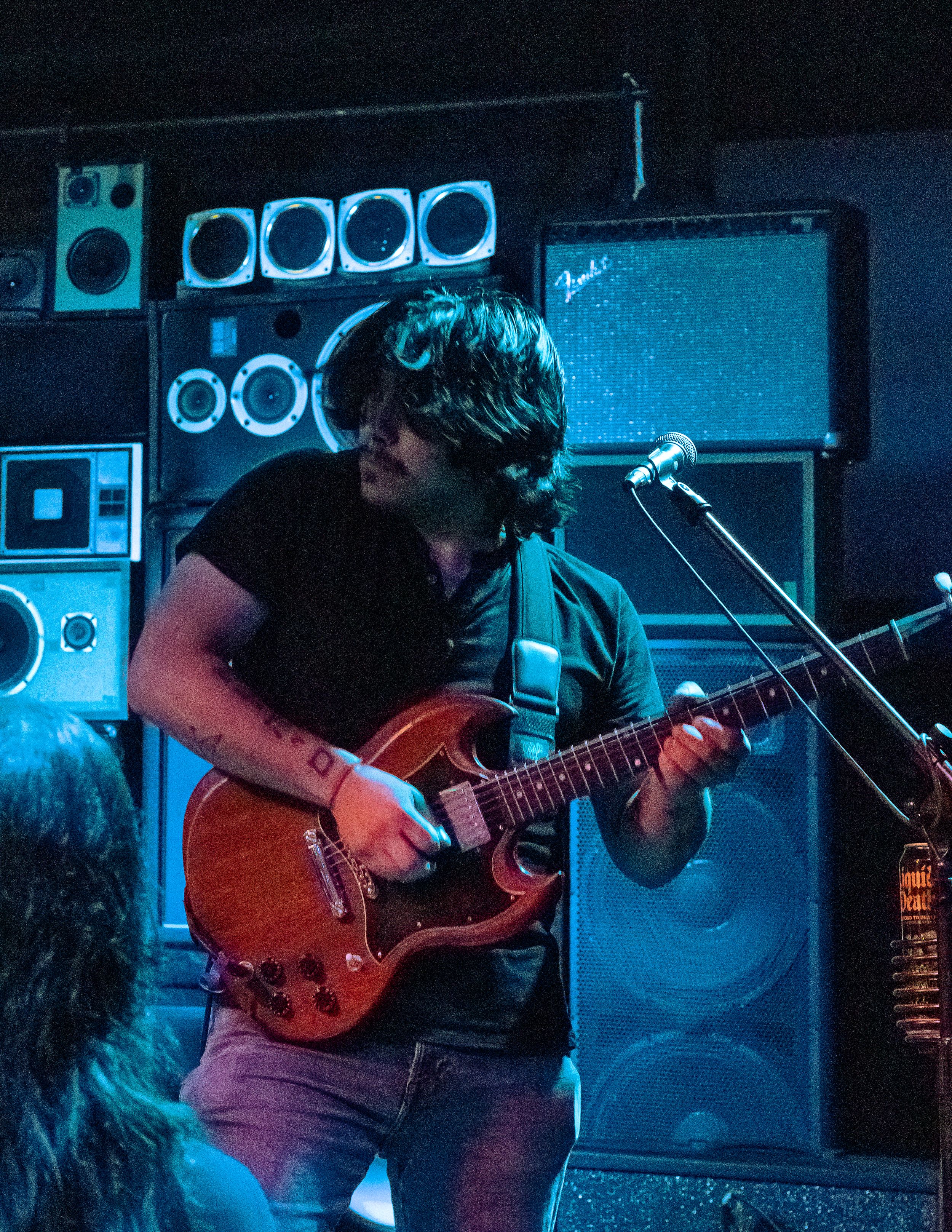 A musician performs on a vintage red electric guitar on stage with blue lighting and speaker cabinets in the background.