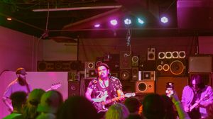 Band performs on stage in a dimly lit venue with purple lighting and stacked speakers in the background.