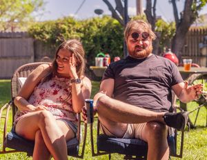 Two people laughing while sitting in outdoor chairs during a casual backyard gathering.
