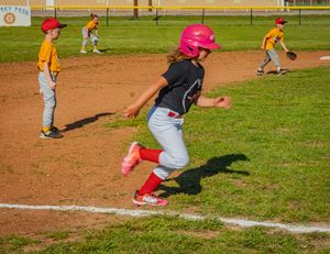 Youth baseball players in action on a dirt field with a runner wearing a pink helmet and cleats taking a lead off base.