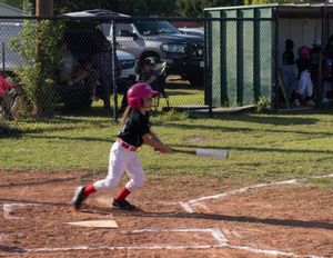 Youth baseball player in pink and black uniform swings bat at home plate during game on dirt field.