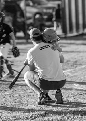 A baseball coach in a white shirt crouches near home plate while holding a child during a game on a sunny day.
