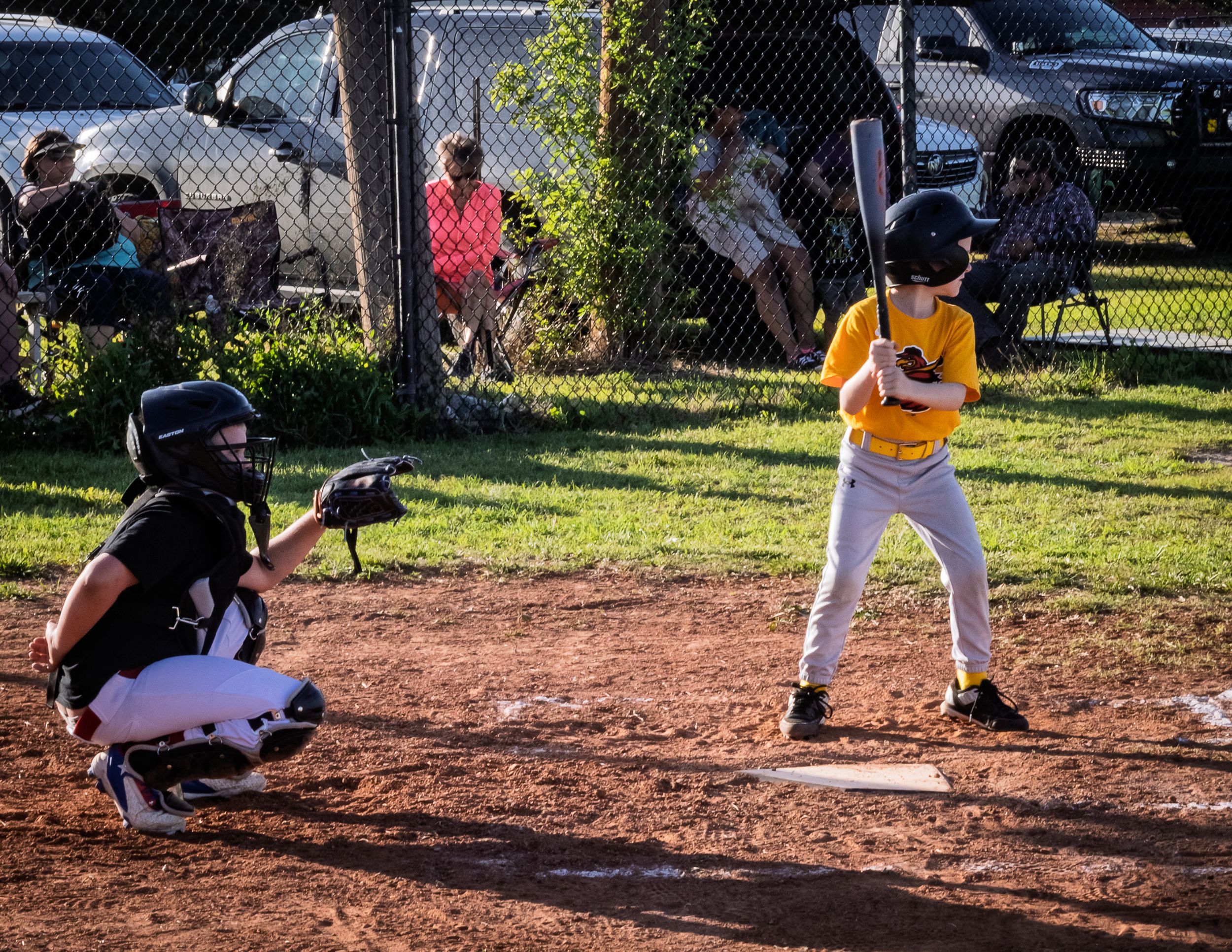Youth baseball player in yellow uniform at bat with catcher crouched behind home plate during game on dirt field.