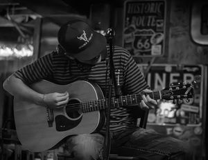 Four black and white images of musicians playing acoustic guitars in a dimly lit venue with vintage signs in the background.