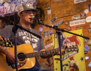 A musician performs with an acoustic guitar at a rustic venue with wooden walls decorated with various signs and stickers.