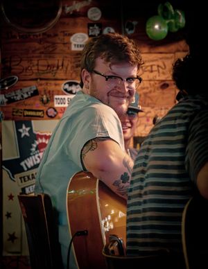 Musician in white t-shirt and glasses sits at wooden bar decorated with signs and memorabilia.