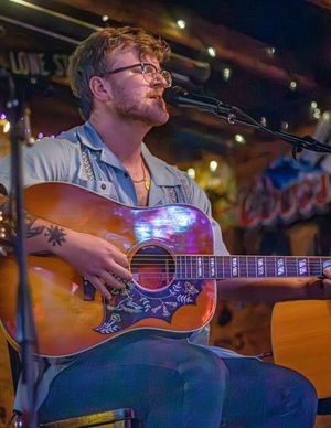 A musician performs with an acoustic guitar in a dimly lit venue with warm lighting and stage ambiance.