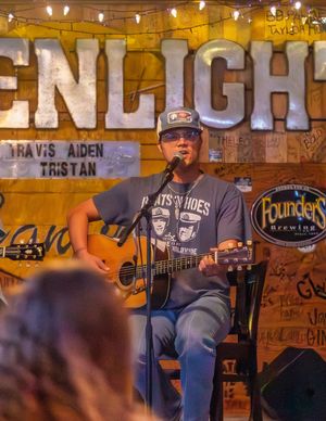 Musicians perform on stage at a dimly lit bar with string lights and handwritten signs on wooden walls in the background.