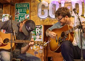 Musicians play acoustic guitars in a rustic venue decorated with vintage Route 66 signs and lighting.