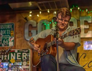A guitarist plays an acoustic guitar in a warmly lit bar or diner setting with neon signs glowing in the background.