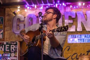 A musician performs passionately on an acoustic guitar under purple stage lights at a venue with rustic wooden walls.