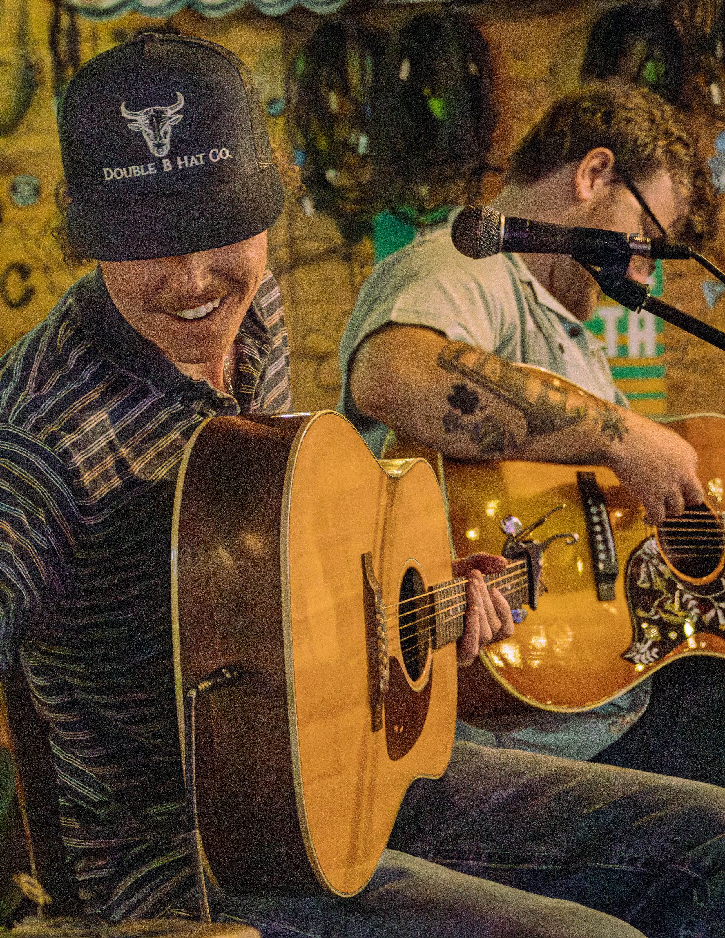 A musician plays an acoustic guitar in a warm-lit venue wearing a striped shirt and baseball cap.