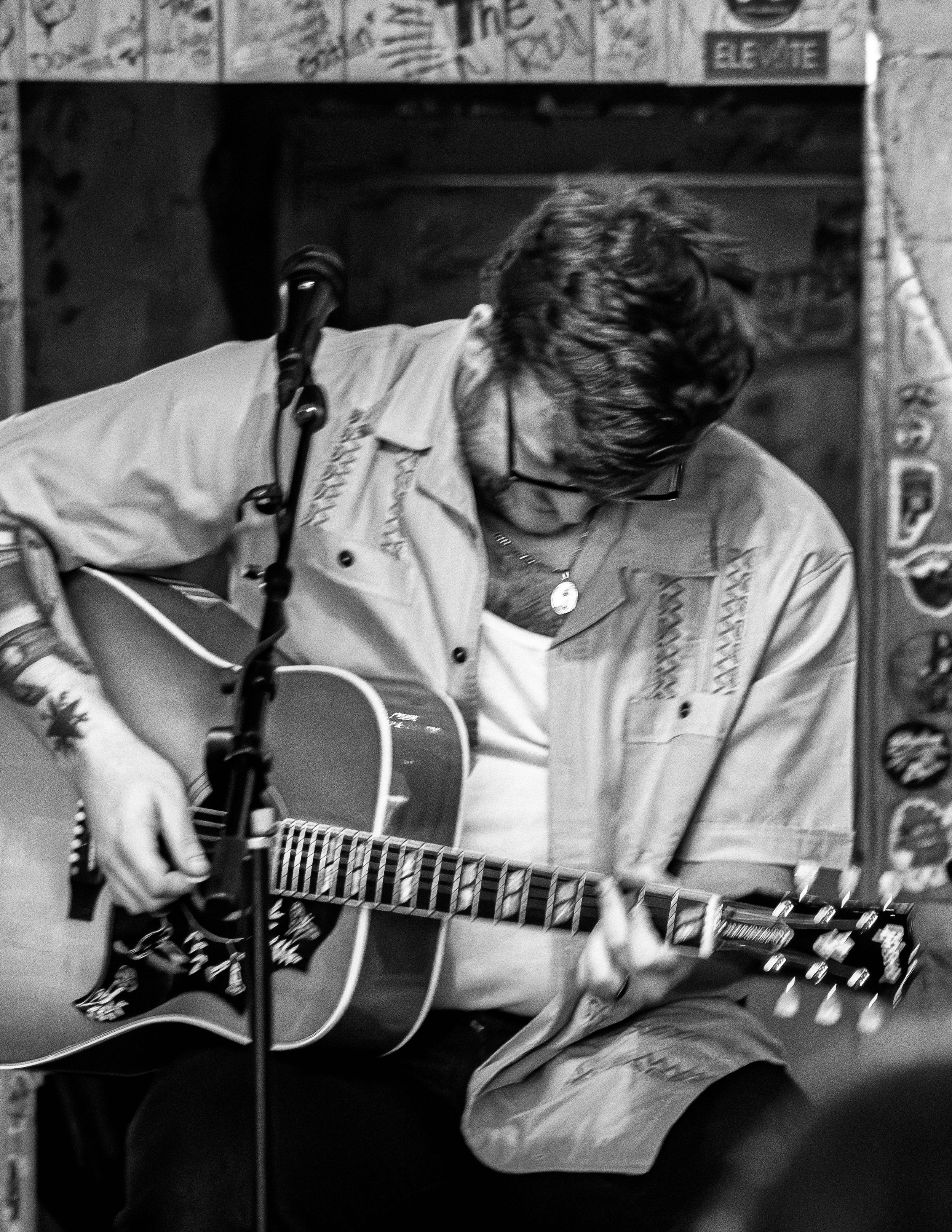 A musician in a light denim jacket plays an acoustic guitar in a black and white photograph.
