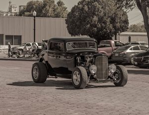 Classic black hot rod car parked in a vintage-style scene with other vehicles in the background.