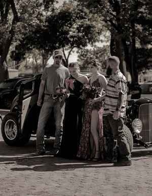 A black and white vintage photo showing a group gathered around a classic car on a brick street.