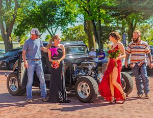 A colorful scene of people in formal attire posing next to a vintage hot rod car on a brick road.