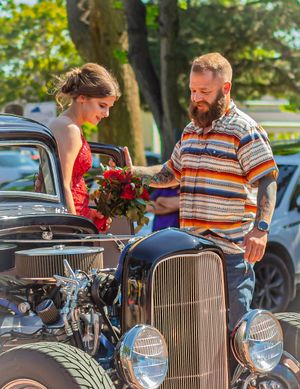 A couple stands beside a vintage hot rod car with an exposed engine while exchanging flowers on a sunny day.