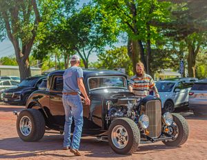 A classic hot rod car enthusiast admires a customized black vintage coupe with exposed engine at an outdoor car show.