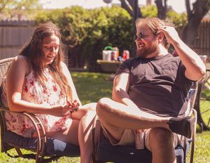 Two people relax together in lawn chairs on a sunny summer day in a backyard garden setting.