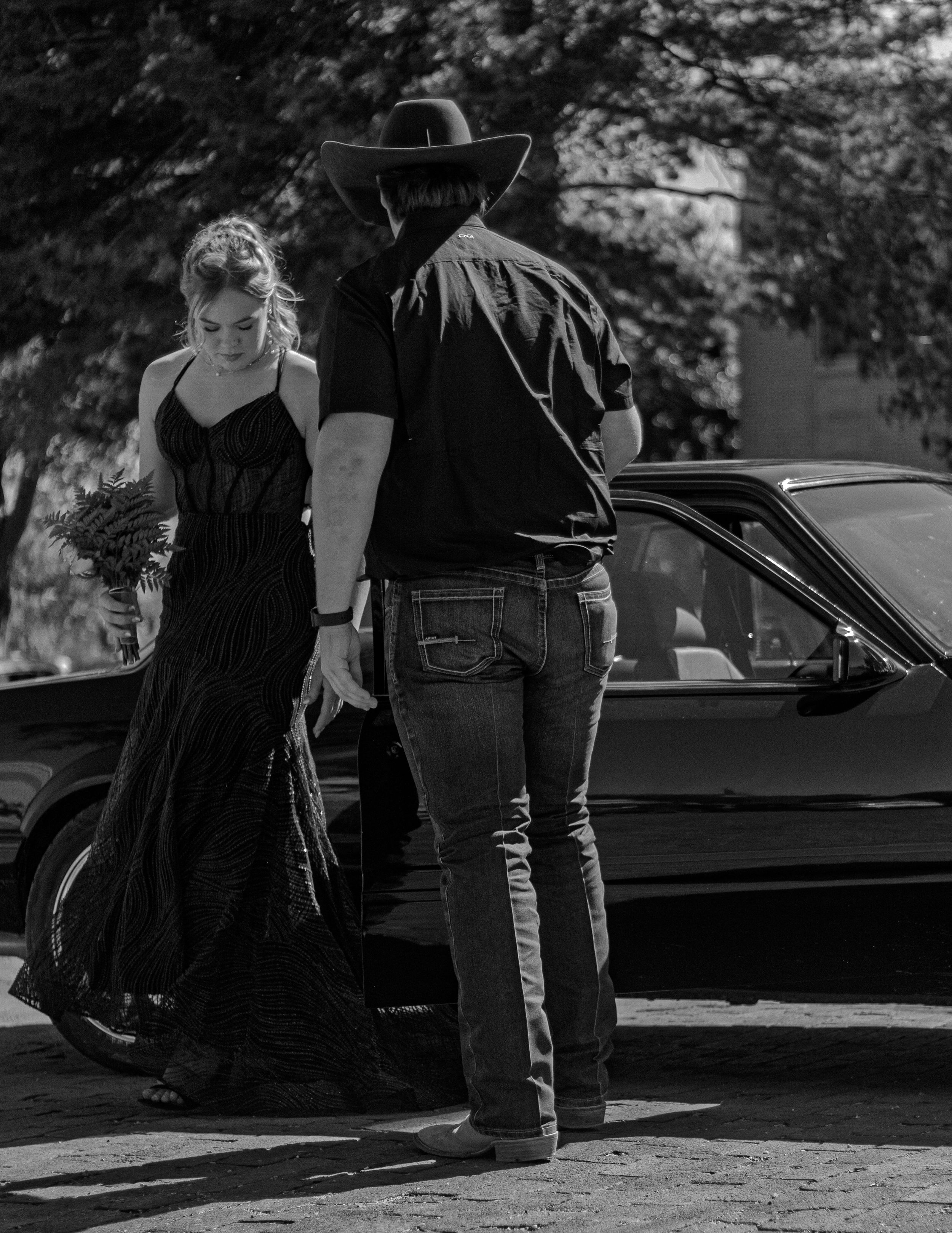 A figure in jeans and cowboy hat stands near a classic car while someone in a flowing black dress walks away.