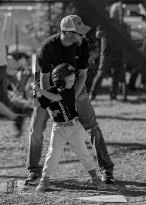 A monochrome photo showing a coach helping a young baseball player practice batting stance at home plate.