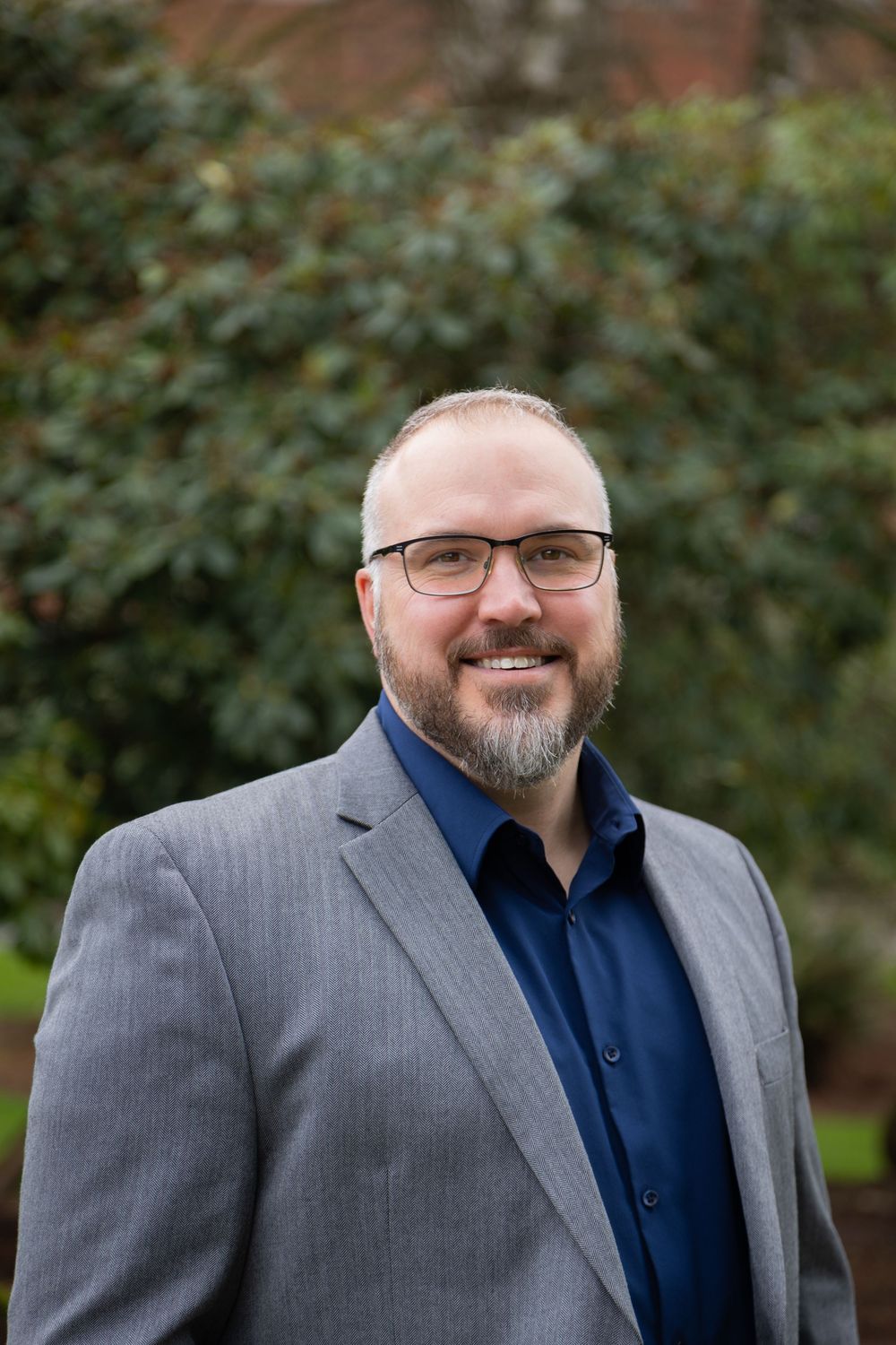 Man in gray blazer and navy shirt with glasses and beard smiling outdoors with green shrubs background.