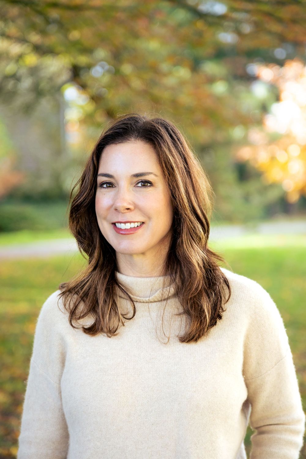 Person wearing cream turtleneck sweater poses outdoors with autumn foliage in background.