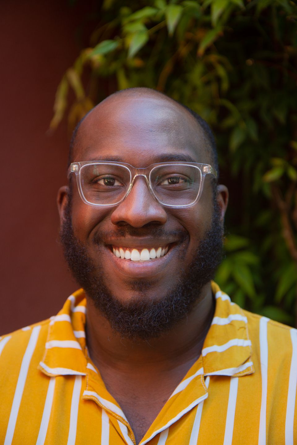 Portrait of someone wearing glasses and a yellow striped shirt, smiling warmly against a green foliage background.