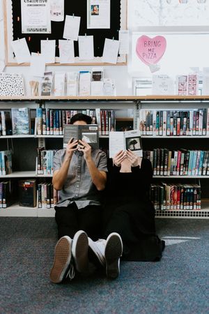 Person sitting on library floor holding books up to cover their face with bookshelves and bulletin board visible.