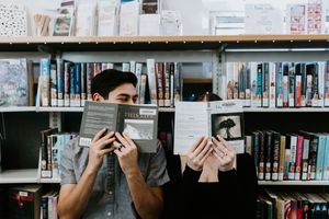 A person peeks through book pages while browsing shelves in a library filled with various publications.