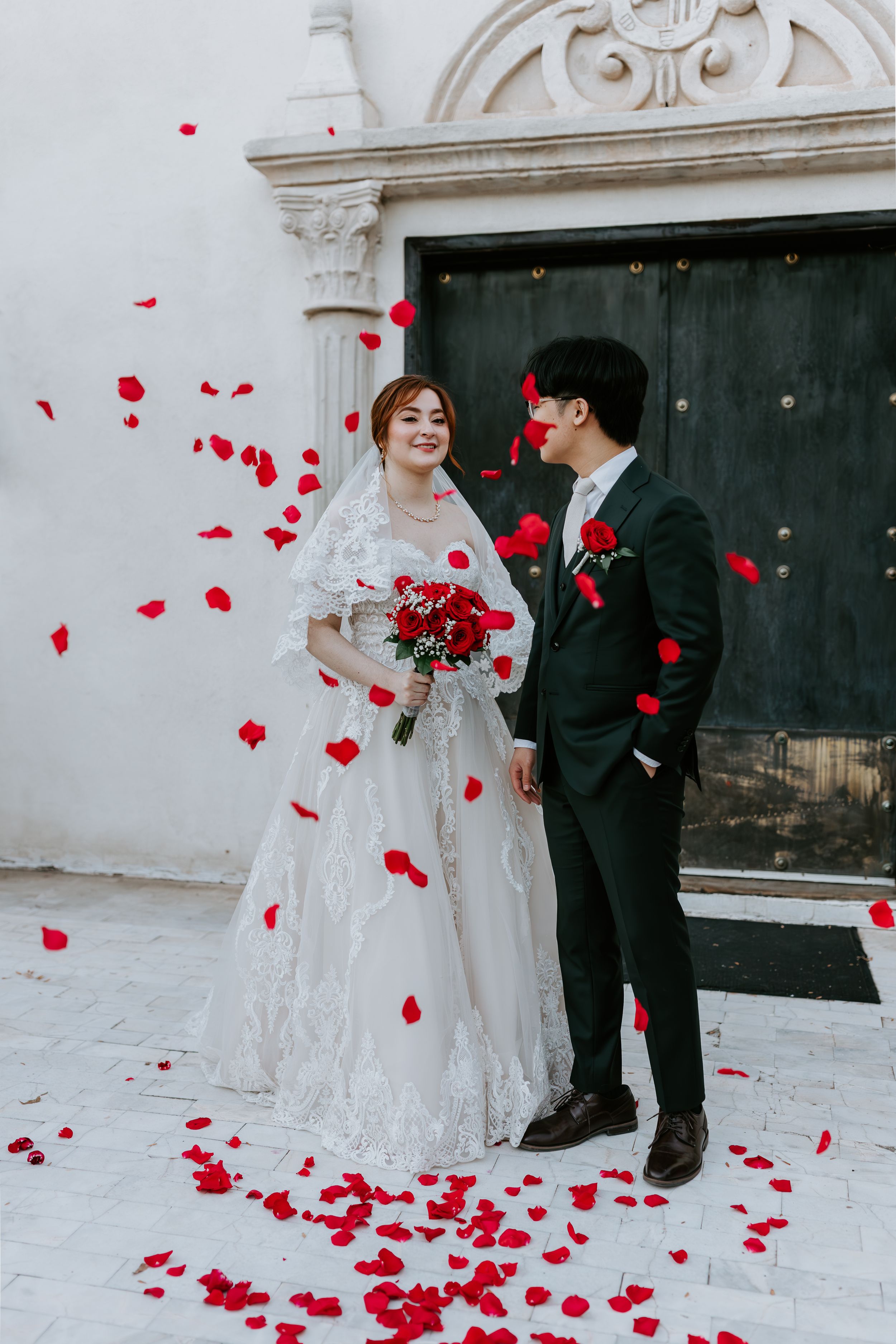 Newlywed couple standing under archway as red rose petals fall around them during wedding celebration.