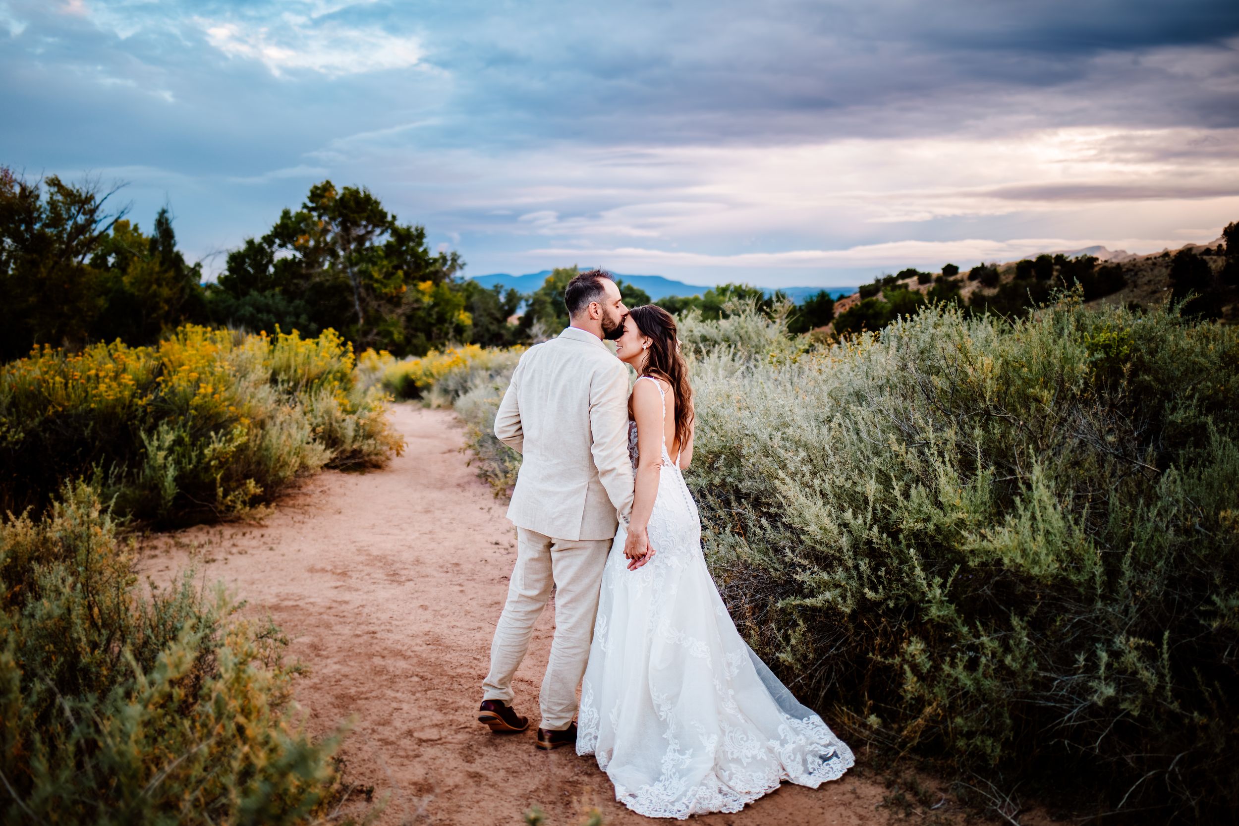 A couple shares a romantic moment on a desert trail surrounded by sagebrush at sunset with dramatic cloudy skies.