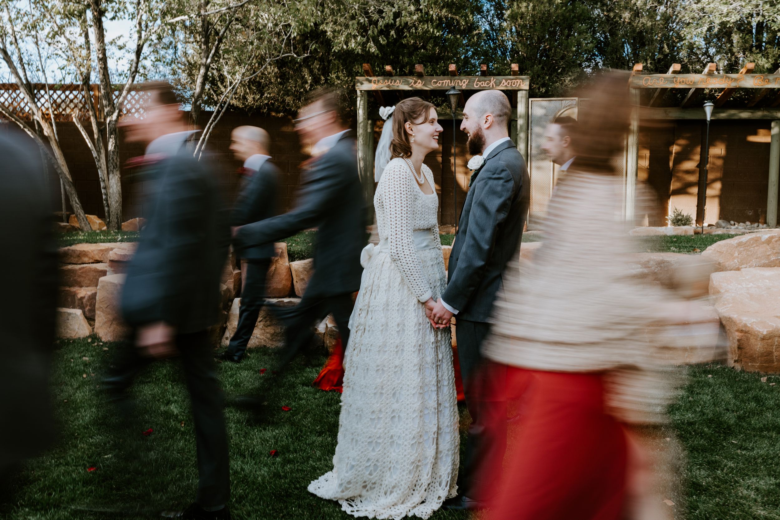 Motion blur captures wedding guests moving past a couple standing together in a garden setting wearing formal attire.