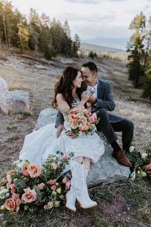 Couple in wedding attire sits on rock in mountains with lace boots and pink floral bouquet arrangements.