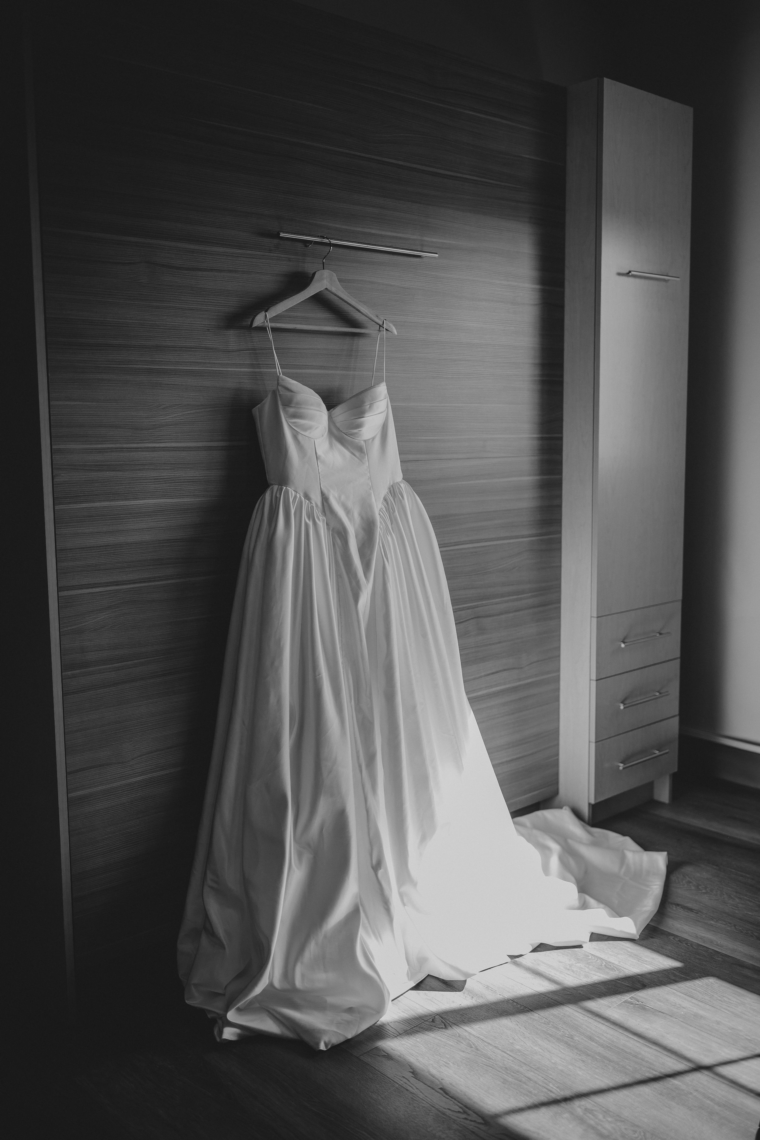 An elegant white wedding dress with a sweetheart neckline and flowy skirt hangs on a wooden hanger against a paneled wall.