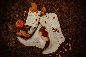 White leather cowboy boots with floral details arranged with dried autumn leaves and orange flowers on dark brown background.