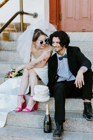 A couple sits on stone steps with a wedding cake, champagne, bright pink heels and sunglasses creating a playful moment.