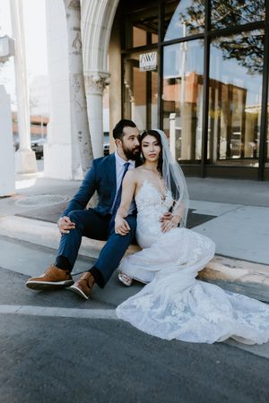 A couple in wedding attire sits casually on steps outside a building with arched windows.