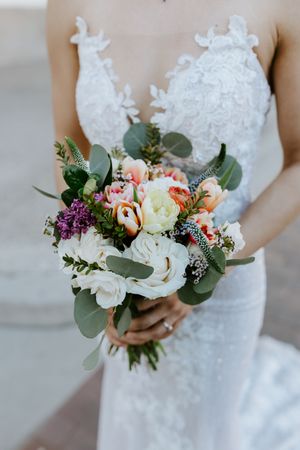 Wedding bouquet with white lace dress features peach ranunculus, purple lilac, and eucalyptus greenery.