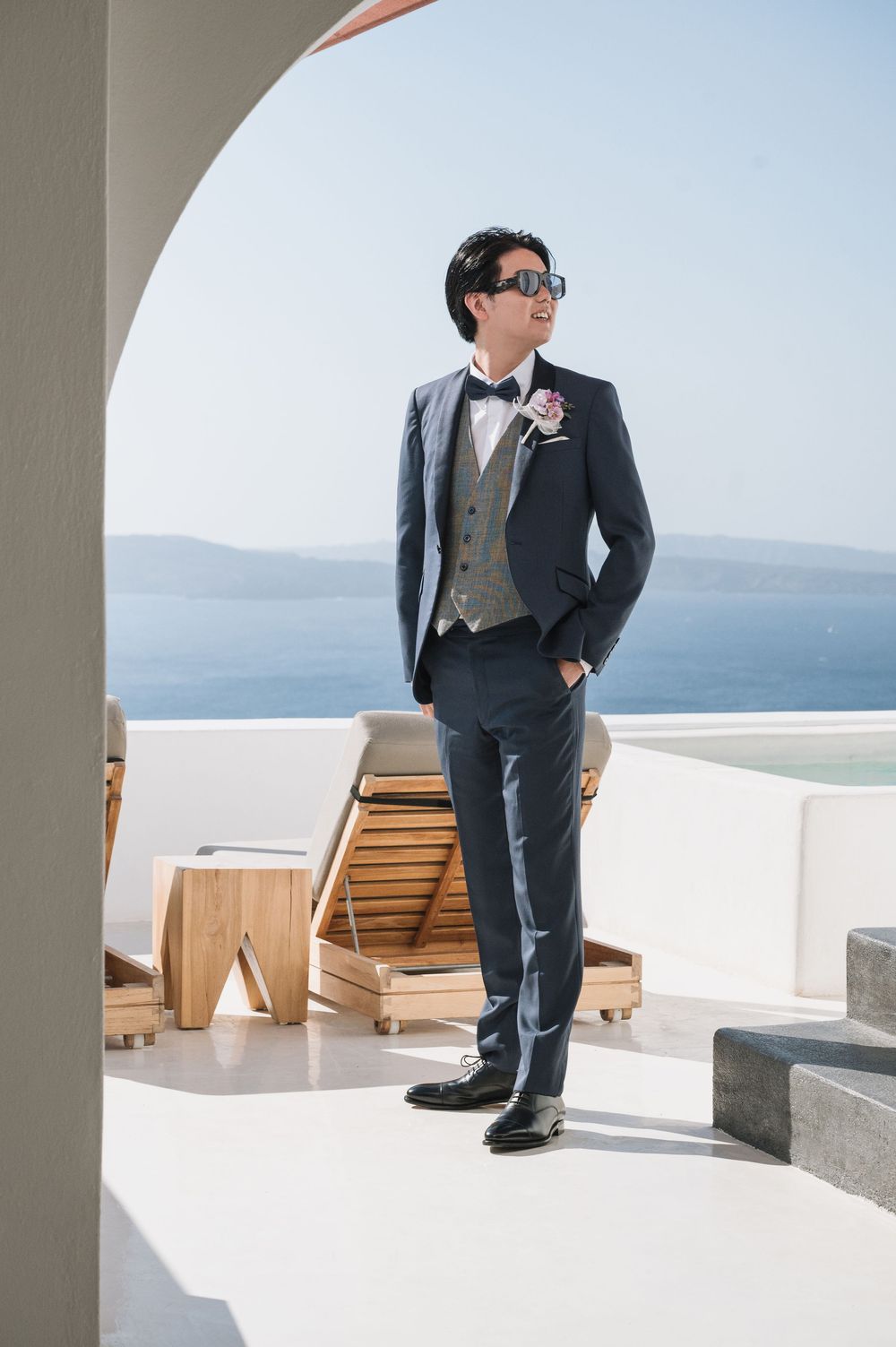 A groom in a formal suit and glasses stands on a white terrace overlooking the Mediterranean Sea.