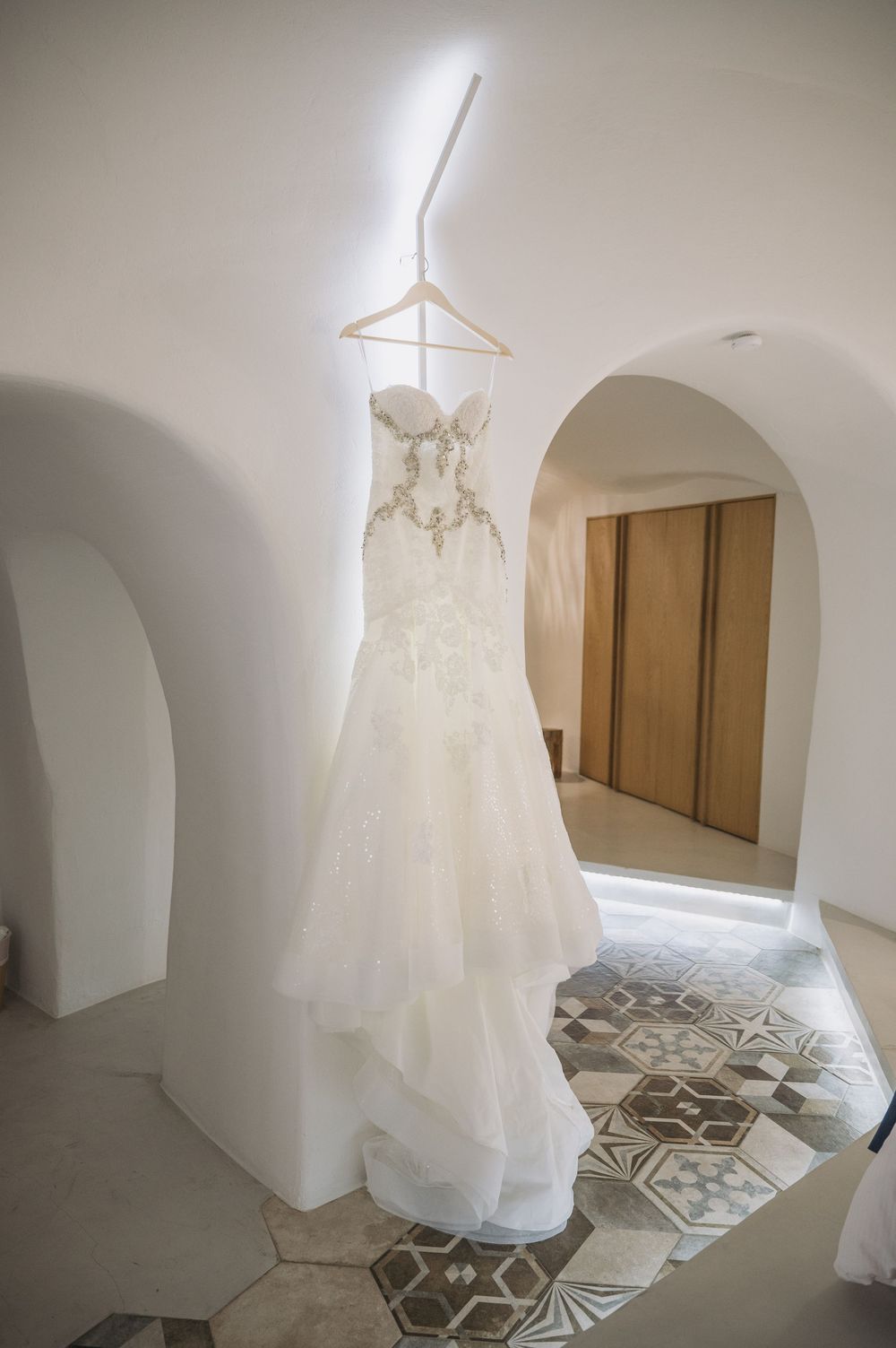 Elegant white lace wedding dress hanging in arched Mediterranean villa hallway with patterned tile floor.