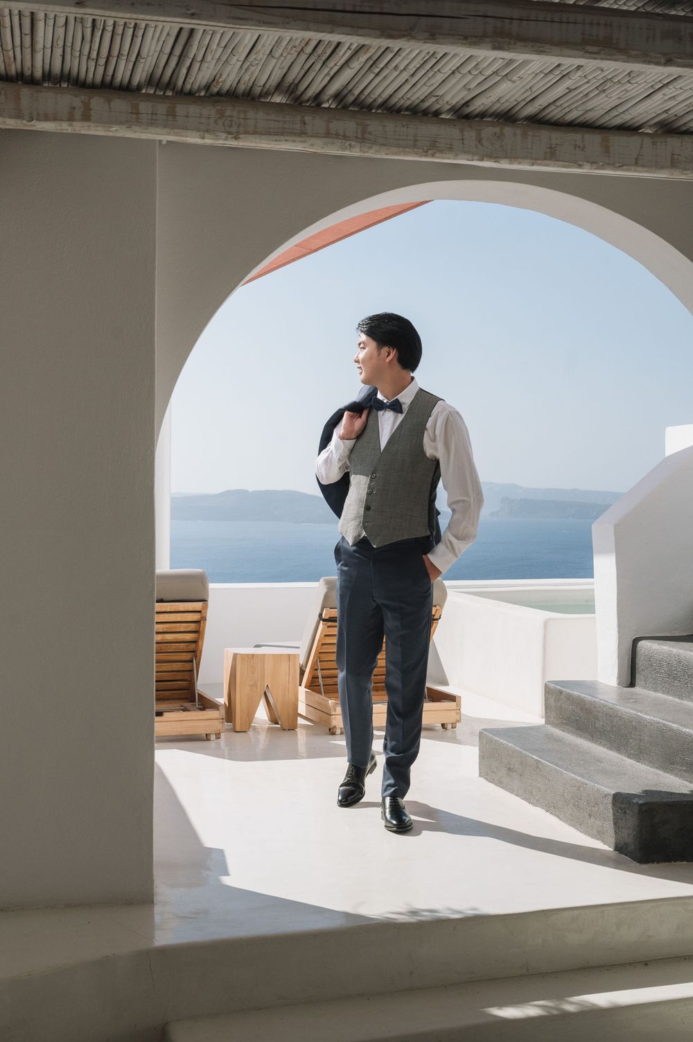 A well-dressed figure in a vest and bowtie stands in a Mediterranean archway overlooking the sea.