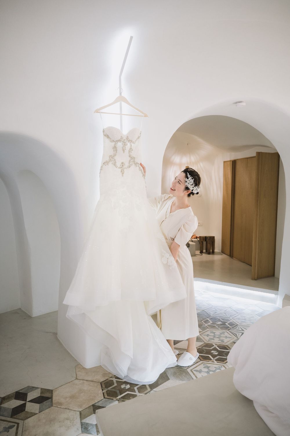 A wedding dress hangs elegantly in a white Mediterranean-style interior with arched doorways and patterned tile floors.