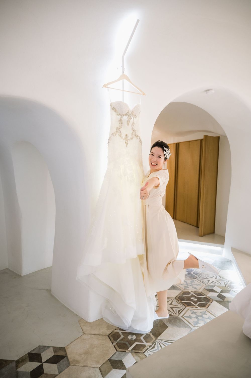 A wedding dress hangs elegantly by a curved archway while someone peeks playfully from behind in an interior space.