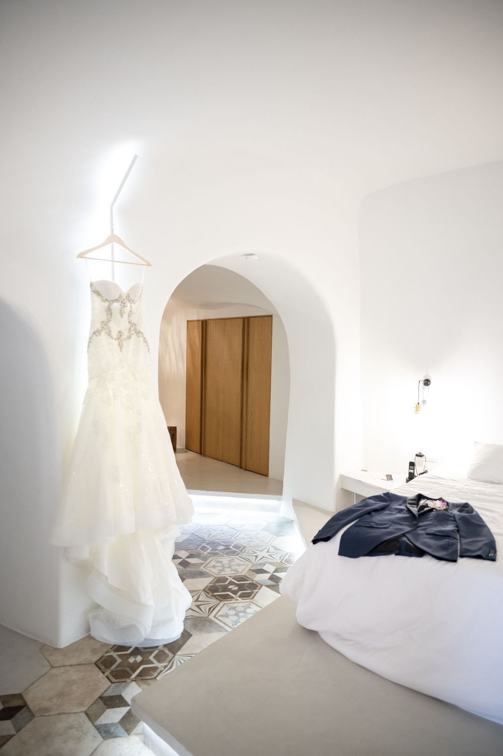 White wedding dress hanging in minimalist white room with arched doorway and patterned tile floor.