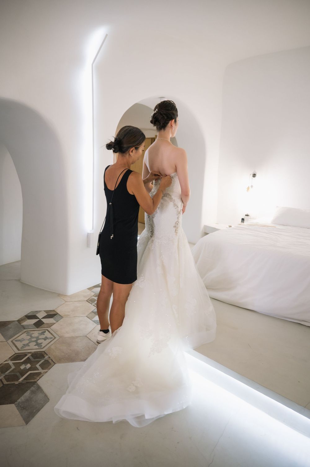 A wedding dress fitting scene in a minimalist white room with curved archways and patterned floor tiles.