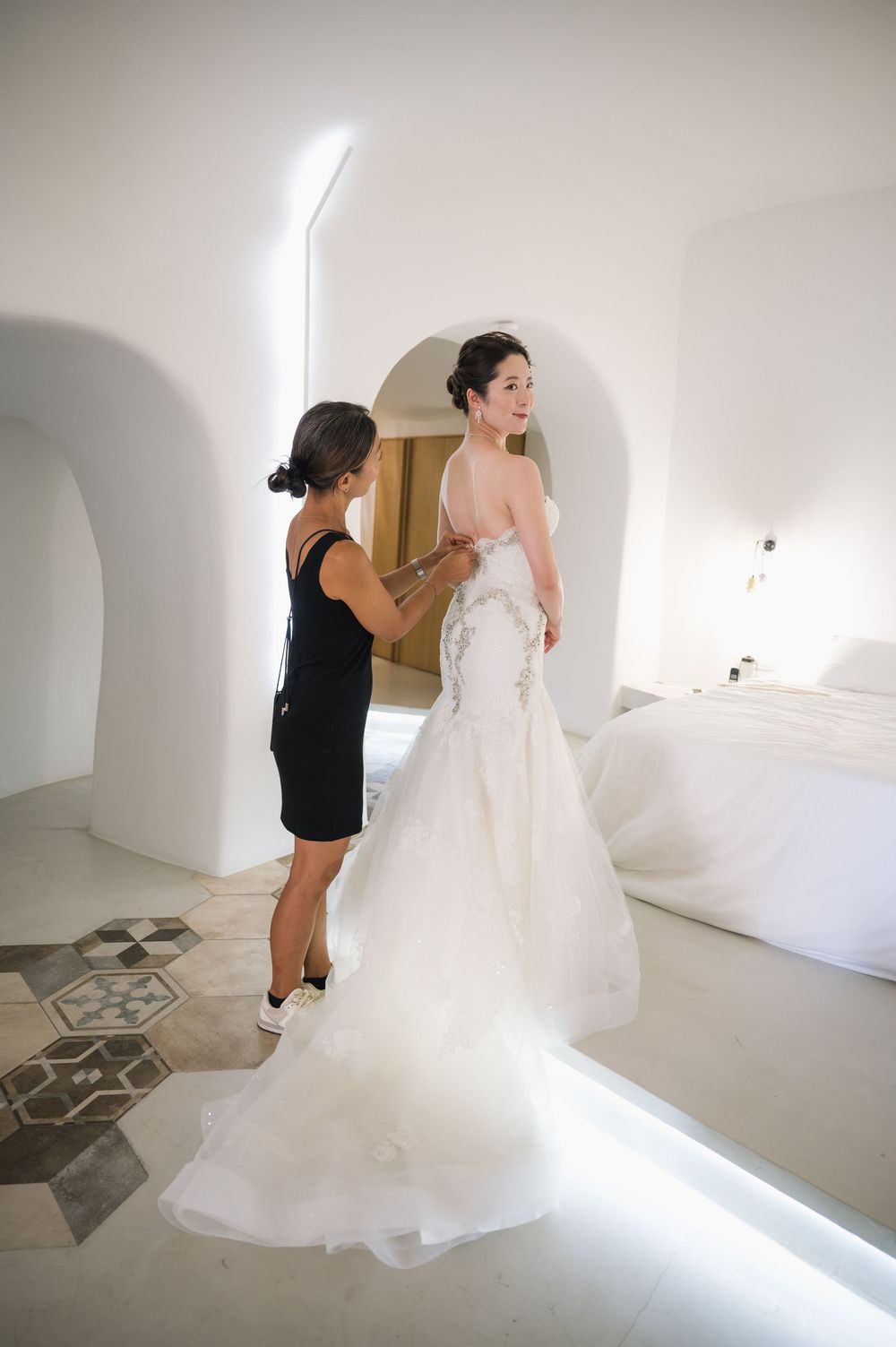 Wedding dress fitting in a bright white Mediterranean-style room with curved archways and tiled floor.